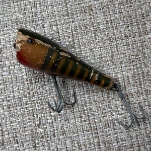 May include: A vintage fishing lure with a tapered wooden body featuring a brown and black striped pattern, a red accent near the front, and a single dark eye. The lure is equipped with two metal treble hooks attached to the underside and tail.