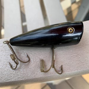 Puede incluir: Un señuelo de pesca negro vintage con un acabado brillante. El señuelo tiene un cuerpo redondeado con un solo ojo y dos anzuelos triples. El ojo es un pequeño círculo dorado. Los anzuelos son de metal y están unidos a la parte inferior del señuelo.