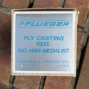 May include: A silver box with blue text that reads "PFLUEGER FLY CASTING REEL NO. 1498 MEDALIST FAYETTEVILLE, ARKANSAS 72701 PT# 8785".