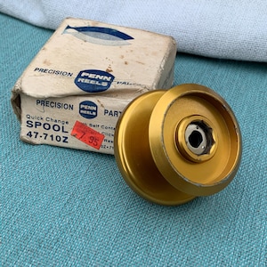 Puede incluir: Un carrete de pesca dorado con un centro hexagonal se muestra junto a su caja original. La caja es beige con texto azul y negro, incluyendo "PENN REELS" y "Quick Change SPOOL 47-710Z". Una etiqueta de precio roja es visible.