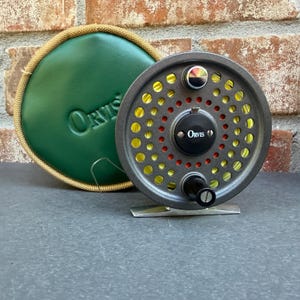 May include: A grey Orvis fly fishing reel with a perforated spool showing yellow line, a black central drag knob, and a black handle. Beside it sits a round, green, zippered protective case with the Orvis logo embossed on the front surface.