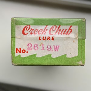 Puede incluir: Una caja verde y blanca con las palabras "Creek Chub Lure" en letras rojas y "No. 2619W" en letras rosas.