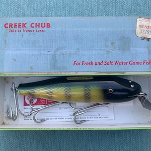 Puede incluir: Un señuelo de pesca Creek Chub en una caja. El señuelo es negro y amarillo con una franja verde. La caja es verde y blanca con el logotipo de Creek Chub y el texto "True-to-Nature Lures".