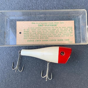 Puede incluir: Un señuelo de pesca blanco y rojo con dos anzuelos triples, en una caja de plástico transparente. El señuelo está etiquetado como "THE 'PLUNKER'" e incluye instrucciones de uso. El señuelo está diseñado para capturar lubinas.