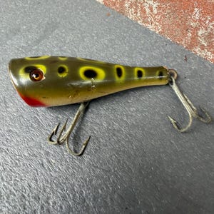 May include: A vintage fishing lure with a smooth, olive-green body featuring yellow-rimmed black spots. The lure has a red-painted chin, a single glass eye, and two silver-toned metal treble hooks attached to the underside and the tail end.