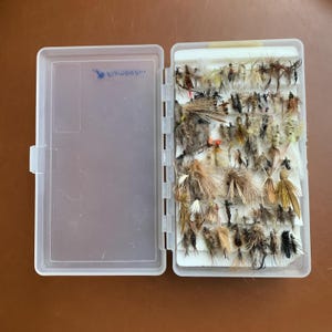 May include: A clear plastic fly fishing box with a white interior. The box is filled with a variety of flies, including dry flies, wet flies, and nymphs. The box has a label on the side that says "Fishwest".