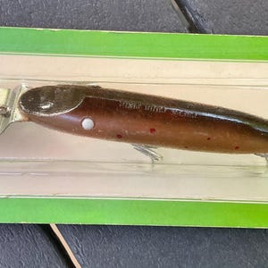 Könnte beinhalten: Ein Vintage-Angelköder in einer transparenten Kunststoffverpackung, vor einem grünen und weißen Kartonhintergrund. Der Köder ist braun mit roten Punkten und einer silbernen Klinge. Die Verpackung trägt den Text "For Fresh and Salt Water Game Fish."