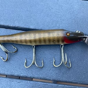 Puede incluir: Un señuelo de pesca con un cuerpo marrón y dorado, con un patrón de escamas. Tiene un acento rojo cerca de la cabeza, una parte superior negra y tres anzuelos triples. El señuelo está diseñado para pescar.