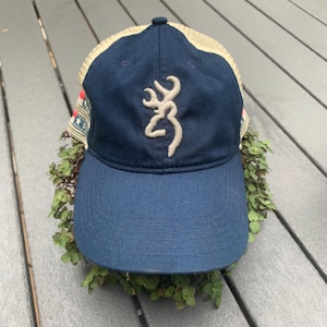 May include: A blue and beige baseball cap with a mesh back. The cap features a white buck logo and a red, white, and blue stripe on the side.