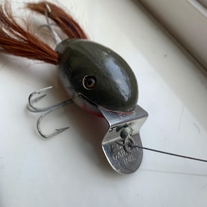 Puede incluir: Un señuelo de pesca de madera verde y rojo con una cola de plumas marrones. El señuelo tiene un anzuelo plateado y una placa plateada en la parte inferior que dice "C.C. GARRETT IND."
