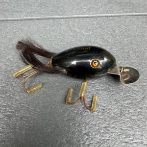 May include: A black fishing lure with the text PINGBAT printed on top. The lure features a single orange eye, a metal diving lip, and dark brown hair attached to the rear. Two metal treble hooks with translucent yellow covers are attached.