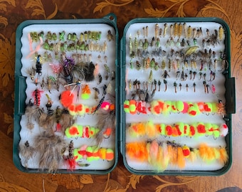 May include: Two green and white plastic boxes filled with colorful fly fishing lures. The lures are arranged in rows and include a variety of colors, sizes, and materials.