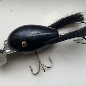 Puede incluir: Un señuelo de pesca de madera negro con un solo ojo y streamers negros parecidos al pelo. El señuelo tiene dos anzuelos triples.