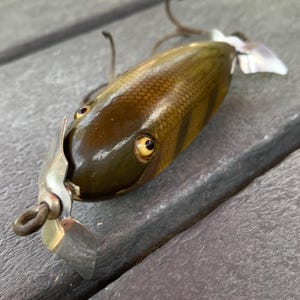 Puede incluir: Un señuelo de pesca vintage con un cuerpo marrón y amarillo, con ojos amarillos y una hélice plateada. El señuelo tiene un patrón de escamas detallado y una pequeña aleta blanca. El anzuelo está unido a la parte delantera del señuelo.