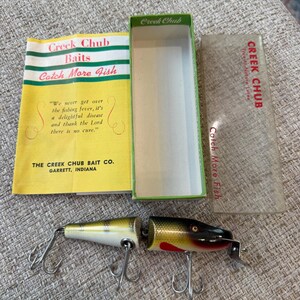 Puede incluir: Un señuelo de pesca vintage Creek Chub con cuerpo segmentado, pintado en amarillo, verde y negro con detalles rojos y anzuelos de metal. Incluye un inserto de papel amarillo con texto negro, una caja de cartón verde y una tapa de plástico transparente.