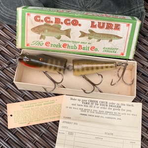 Puede incluir: Un señuelo de pesca vintage Creek Chub Bait Co. en una caja. El señuelo es un pez de madera articulado, marrón y verde con ojos rojos. La caja es verde y blanca con el logotipo de la empresa y las palabras "C.C.B.CO. LURE" y "The Creek Chub Bait Co."