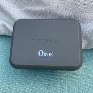 Może przedstawiać: Prostokątne, szare etui z zaokrąglonymi rogami. U góry wydrukowano tekst "C&F DESIGN CONCEPT & FORM BY K.YONENOI", a poniżej wyśrodkowano logo "Orvis". Etui znajduje się na teksturowanej powierzchni.