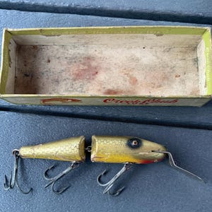 Puede incluir: Señuelo de pesca Creek Chub vintage en amarillo y dorado, con diseño dividido y tres anzuelos triples. El señuelo está junto a su caja de cartón original y desgastada, que tiene las palabras "Creek Chub" impresas.