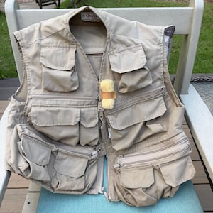 Pode incluir: Um colete utilitário bege da marca Orvis com vários bolsos com fecho e aba na parte frontal. A peça inclui um pequeno emblema felpudo de cor creme fixado na zona do peito, um fecho central e uma etiqueta da marca na gola.