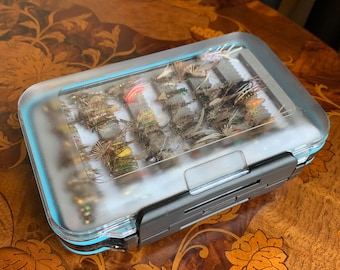 Puede incluir: Una caja de pesca con mosca de plástico transparente con un cierre negro. La caja está llena de moscas de colores.