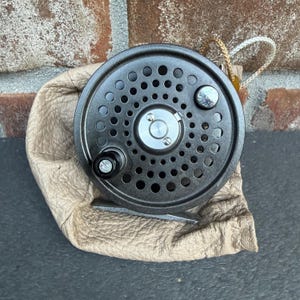 May include: A dark grey metal fly fishing reel with a circular perforated frame, a central silver cap, and a black handle knob. The reel is resting on a beige textured leather pouch with a braided cord drawstring visible at the top right corner.
