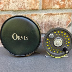 May include: A round black zippered storage case with the word Orvis printed in white, positioned next to a grey fly fishing reel featuring a perforated frame and bright yellow fishing line wound around the central spool mechanism.