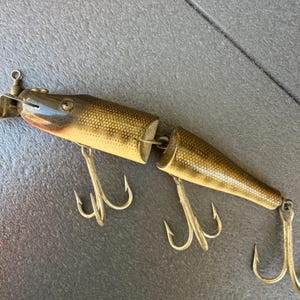 Puede incluir: Un señuelo de pesca vintage con un cuerpo segmentado y tres anzuelos triples. El señuelo tiene un acabado metálico con un patrón de escamas y un acento rojo cerca de la cabeza. El señuelo está diseñado para atraer peces.