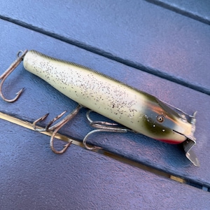 Puede incluir: Un señuelo de pesca vintage con un cuerpo color crema salpicado de puntos negros. El señuelo tiene una cabeza verde y marrón con boca roja y aleta metálica plateada. Tres anzuelos oxidados están unidos.
