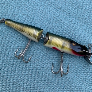 Puede incluir: Un señuelo de pesca vintage con un cuerpo segmentado, pintado en tonos verdes, amarillos y negros, con detalles rojos. Cuenta con dos juegos de anzuelos triples y una pequeña aleta de cola metálica. El señuelo está diseñado para imitar a un pez pequeño.