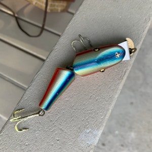 Puede incluir: Un señuelo de pesca de madera con un esquema de pintura azul, dorado y rojo. El señuelo tiene dos anzuelos triples y un giratorio.