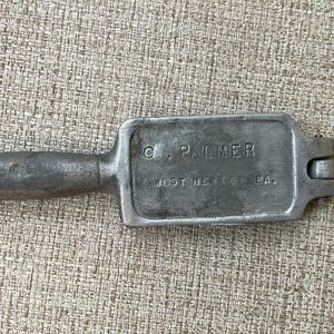 May include: A silver-coloured metal kitchen tool with a rectangular head and a rounded handle. The surface features embossed text reading C. PALMER and WEST NEWTON PA. The tool has a hinged mechanism on one end and a textured, weathered metallic finish.