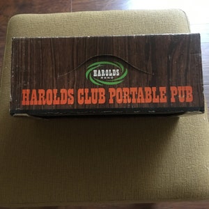 May include: A brown cardboard box with a woodgrain pattern. The box has a green and white logo that says "HAROLDS RENO" and the text "HAROLDS CLUB PORTABLE PUB" in orange letters.