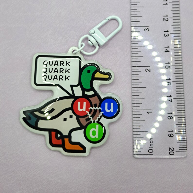 Op de afbeelding: Een witte eend sleutelhanger met een groene kop en een rode, blauwe en groene cirkel op zijn borst. De cirkels hebben de letters "u", "u" en "d" erop. De eend houdt een spreekballon vast waarop "QUARK QUARK QUARK" staat.