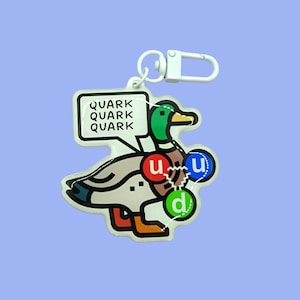 Op de afbeelding: Witte sleutelhanger met een cartoon eend en de woorden "Quark Quark Quark" in een spraakballon. De eend heeft rode, blauwe en groene cirkels op zijn lichaam met de letters "u", "d" en "u" erin.