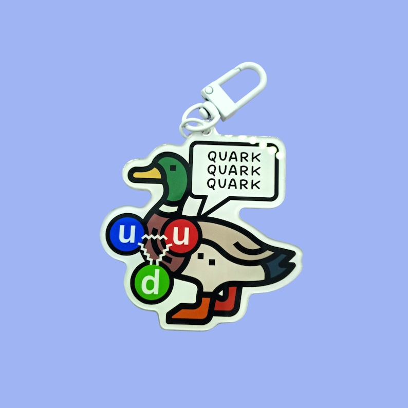 Op de afbeelding: Een witte sleutelhanger met een cartoon eend en de woorden "Quark Quark Quark" op een witte spraakballon. De eend heeft een blauwe, rode en groene cirkel op zijn lichaam.