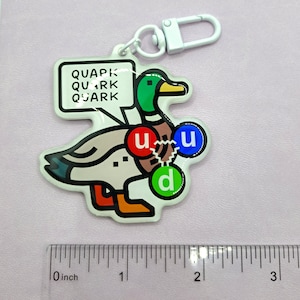 Op de afbeelding: Een witte eend sleutelhanger met een groene kop en een spraakballon die "QUAPK QUARK QUARK" zegt. De eend heeft drie gekleurde cirkels op zijn lichaam met de letters "u", "u" en "d" erin.