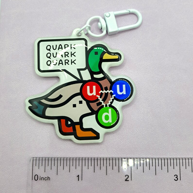 Op de afbeelding: Een witte eend sleutelhanger met een groene kop en een spraakballon die "QUAPK QUARK QUARK" zegt. De eend heeft drie gekleurde cirkels op zijn lichaam met de letters "u", "u" en "d" erin.