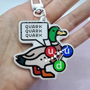 Op de afbeelding: Een witte eend sleutelhanger met de tekst "Quark Quark Quark" in een spreekballon erboven. De eend heeft drie gekleurde cirkels op zijn lichaam, met de labels "u", "u" en "d".