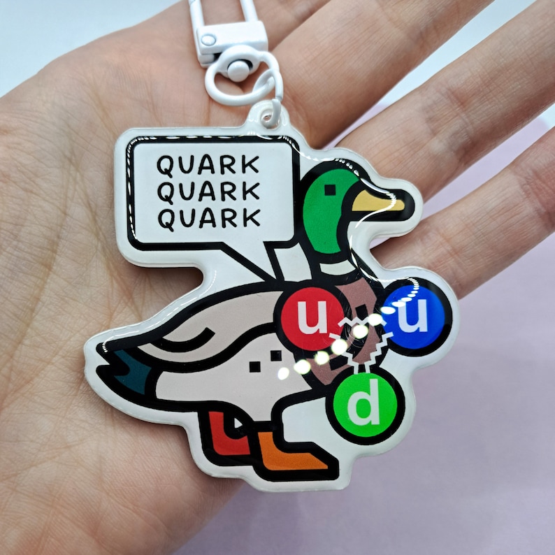 Op de afbeelding: Een witte eend sleutelhanger met de tekst "Quark Quark Quark" in een spreekballon erboven. De eend heeft drie gekleurde cirkels op zijn lichaam, met de labels "u", "u" en "d".