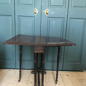 May include: A dark brown, antique drop-leaf table with a rectangular top and four slender, turned legs. The table has a decorative apron and is set against a teal-colored door. The table is approximately 76 cm long when fully extended.