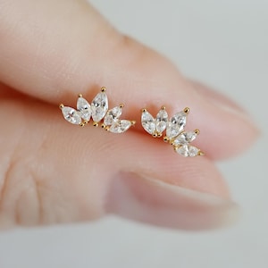 May include: A pair of gold-toned stud earrings, each featuring a cluster of marquise-cut, clear cubic zirconia stones. The earrings are designed in a floral or leaf-like pattern, with the stones arranged to create a delicate, sparkling effect. The earrings are shown on a light background.