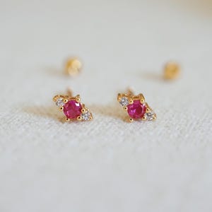 Puede incluir: Un par de pendientes de oro, cada uno con una vibrante gema roja en el centro, flanqueada por piedras de acento claras más pequeñas. Los pendientes están colocados sobre un fondo suave y neutro, resaltando su delicado diseño.