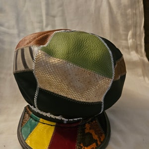 May include: A patchwork cap featuring a variety of textures and colors. The cap's crown is composed of leather and faux animal print panels in shades of brown, green, and black. The brim is a patchwork of red, yellow, and green leather.