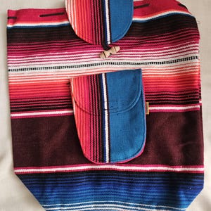 May include: A woven fabric backpack featuring a multi-colored striped pattern in shades of red, pink, white, dark brown, and blue. The bag includes a top flap with a wooden toggle closure and a smaller front pocket with a rounded flap closure.