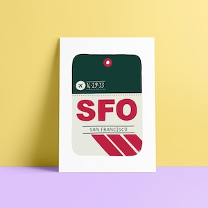 Könnte beinhalten: Weißer Druck mit einem stilisierten Reiseanhänger-Design. Der Anhänger ist dunkelgrün und hellgrau, mit den Buchstaben "SFO" in Rot und den Worten "San Francisco" darunter. Ein kleines Flugzeugsymbol ist sichtbar.