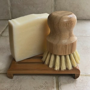 May include: A wooden dish brush with natural bristles and a wooden soap dish with a bar of white soap. The soap dish and brush are on a bamboo soap dish.