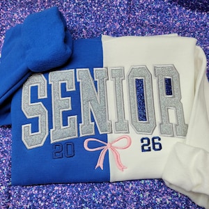May include: A blue and white split-color sweatshirt with the word "SENIOR" in silver glitter lettering. The year "26" is embroidered in blue, and "20" is embroidered in black. A pink bow is embroidered below the text.