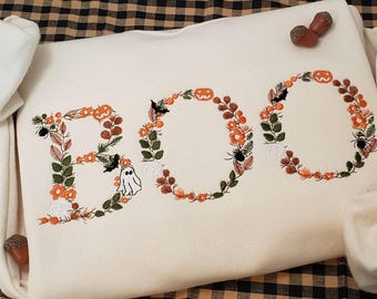 May include: A white sweatshirt with the word "BOO" embroidered in orange, green, and black. The letters are made of wreaths with pumpkins, bats, spiders, and flowers.