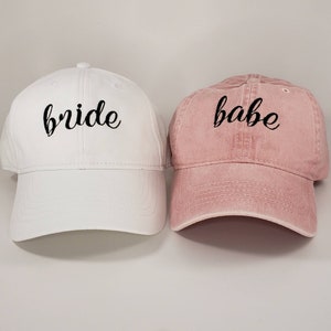 May include: Two baseball caps, one white with the word "bride" embroidered in black, and one pink with the word "babe" embroidered in black.