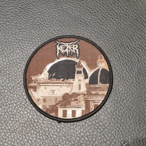 May include: Round patch with a brown and black design featuring a cityscape with the word "KETER" in a stylized font. The patch has a black border and is set against a textured, dark gray surface.